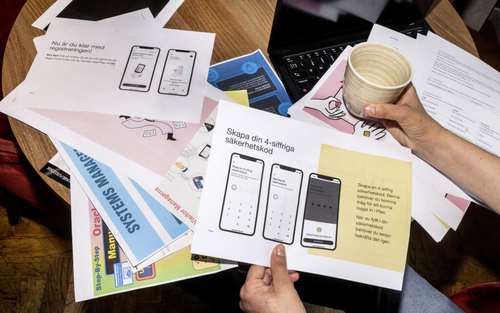 Bildtitel: "Manualparty". Bilden visar ett runt bord som är täckt av utskrivna bilder som ligger huller om buller. På bilderna finns information kring säkerhetskoder och registreringar. Under pappershögen finns en bärbar dator. Två händer är synliga ovan pappershögen, varav den ena håller en kaffekopp.